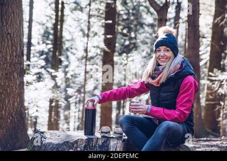 A smiling female tourist takes a break for hot tea from a thermos and a meal on the mountain trail. Stock Photo