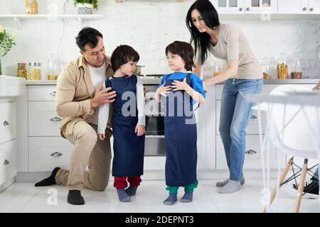 Hispanic parents puttting on aprons on two little boys, twins while cooking dinner in the kitchen at home together. Happy family, children, cooking concept Stock Photo