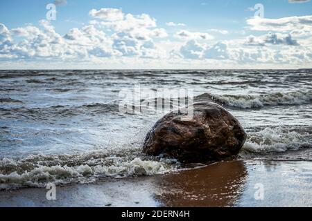 The wave beats against a large stone on the seashore, a clear day, light clouds Stock Photo