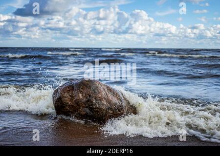 The wave beats against a large stone on the seashore, a clear day, light clouds Stock Photo