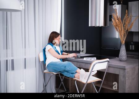 Young woman working remotly from her dining table. Home kitchen in the background. Stock Photo