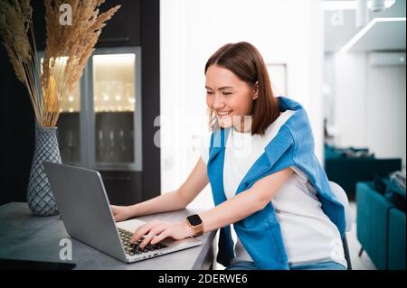Smiling woman sitting in the kitchen and using the laptop. Working from home in quarantine lockdown. Social distancing Self Isolation. Stock Photo