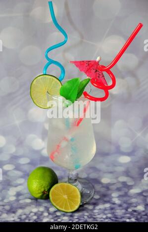 A glass of lime juice with ice, straws, cocktail umbrellas and green citrus fruits on a gray shiny background. Close-up. Stock Photo