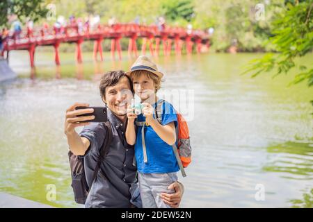 Caucasian Dad and son travelers on background of Red Bridge in public park garden with trees and reflection in the middle of Hoan Kiem Lake in Stock Photo