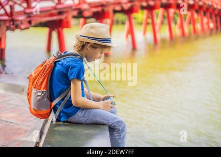 Caucasian boy tourist on background of Red Bridge in public park garden with trees and reflection in the middle of Hoan Kiem Lake in Downtown Hanoi Stock Photo