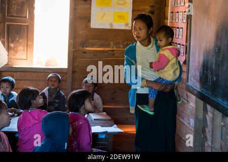 Teacher holding her baby by blackboard inside elementary school classroom with pupils in Kayan village, near Loikaw, Myanmar Stock Photo