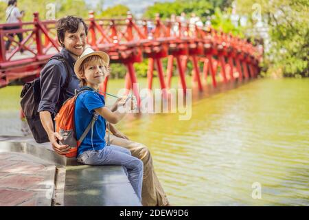 Caucasian Dad and son travelers on background of Red Bridge in public park garden with trees and reflection in the middle of Hoan Kiem Lake in Stock Photo