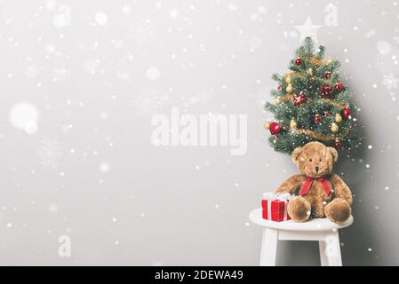 Lovely Christmas holiday background with red gift boxes, teddy bear, small decorated Pine tree and falling snow in white gray room Stock Photo