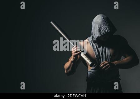 Dangerous man in the mask with baseball bat ready for fight. An aggressive muscular hooded man is holding a baseball bat. Stock Photo