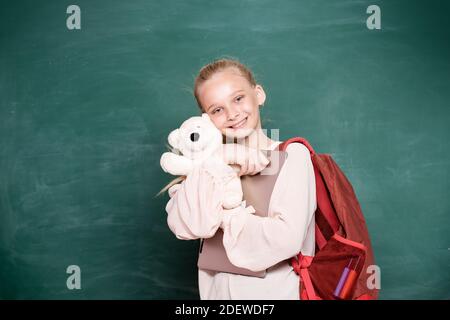 Cute teen student. Attractive little girl holding backpack and teddy while standing against blackboard. Portrait of modern happy teenager school girl Stock Photo