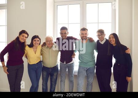 Team of smiling diverse people standing by the window, hugging and looking at camera Stock Photo