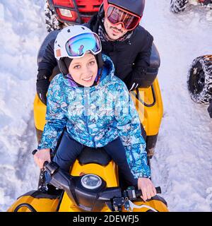 Top view of joyful young woman and man in helmets and goggles driving all-terrain vehicle through snow. Female and male riders looking at camera and smiling while sitting on quad bike. Stock Photo