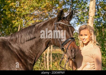 Side view, portrait. Female in casual clothes stands in the forest with her horse. In the autumn sun. Stock Photo