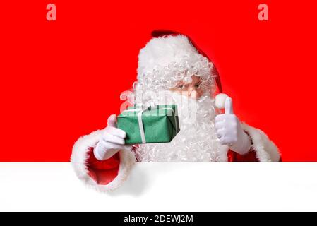 Santa Claus holding green gift box in hand while standing behind the blank white board. copy space. Stock Photo
