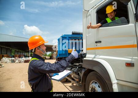 Asian foreman with safety hats and safety vest is carrying a car inspection document in the parking with truck drivers,Concept of planning work day. R Stock Photo