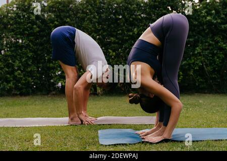 Side view of man and woman doing Standing Forward Bend on mats while practicing yoga in backyard on summer day Stock Photo