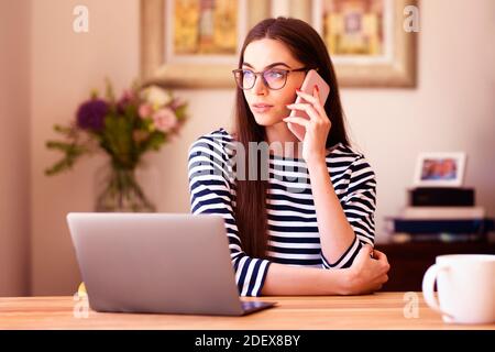 Shot of attractive young businesswoman talking with somebody on her mobile phone while working on laptop at home. Home office. Stock Photo