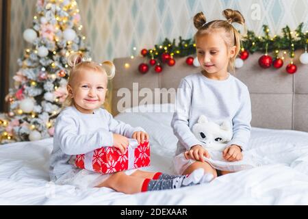 Two cute girls sisters with Christmas presents sit on bed at home in Christmas decorated room Stock Photo