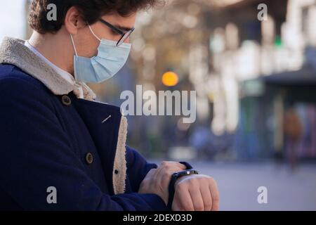 A recognizable young man wearing a mask looks at his watch on a winter day in the city center. Man is in a hurry because he is late for work. Stock Photo