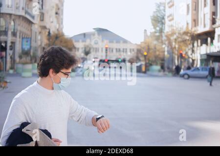 Panoramic recognizable young man wearing a mask with a coat in his hands. He looks at his watch on a winter day in the city center. Man is in a hurry. Stock Photo