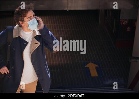 Recognizable young busy man with a mask talking on the phone outside the underground in the city center. Working and business day 2021. Stock Photo