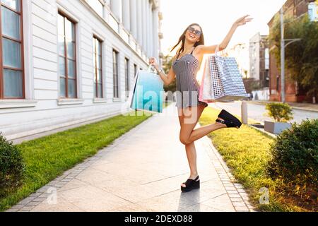 Young attractive girl in sunglasses, enjoys a successful shopping, on the background of a beautiful sunset, with bags in her hands Stock Photo