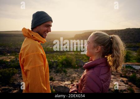 Laughing young male and female friends looking at each other enjoying sunrise on mountain edge Stock Photo