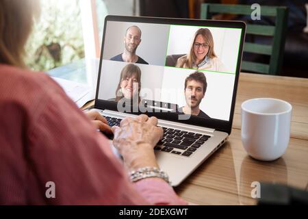 View over shoulder of senior woman talking by video call with a group of young. Laptop screen with smiling people.Elderly grandmother over 60s enjoy v Stock Photo