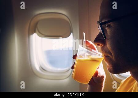 Young passenger enjoying dring during flight. Man holding cocktail against airplane window. Stock Photo