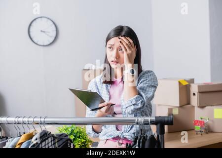 Upset asian volunteer holding clipboard with pen near hanging rack with clothes on blurred foreground in charity center Stock Photo