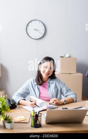 Smiling asian volunteer looking at camera while leafing through notebook at desk with devices and plants in charity center Stock Photo
