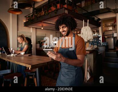 Male waiter with curly hair working in cafe wearing apron with using digital tablet with female working in background on laptop Stock Photo