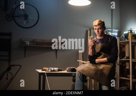 a young carpenter writes a message on his smartphone in a carpenter's workshop while sitting on his Desk at lunchtime Stock Photo