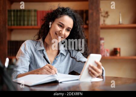 Woman sitting at desk, using smartphone and writing in notebook Stock Photo