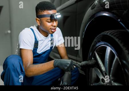 Car washing and detailing workshop. African man worker in protective overalls and rubber gloves, washing car alloy wheel on a car wash, using special Stock Photo