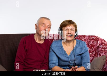 The older woman communicates online using large headphones and a microphone. The spouse is peeping. Stock Photo