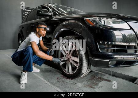 Handsome young dark skinned male worker in uniform, cleaning the wheel rim of modern black crossover car by special brush. Car wash concept and Stock Photo