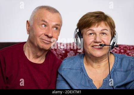 A portrait of a senior woman near husband communicates online using big headphones and microphone.  Stock Photo