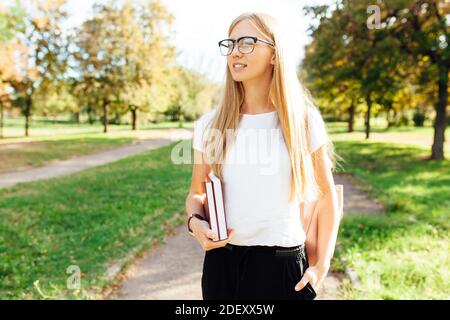 Portrait of a beautiful student girl with glasses, walking in the Park, holding books in her hands, rest after class Stock Photo