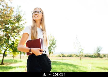 Portrait of a beautiful student girl with glasses, walking in the Park, holding books in her hands, rest after class Stock Photo