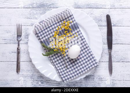 Easter table setting. White egg, napkin on a plate, mimosa flowers, fork, knife on a wooden table. View from above Stock Photo