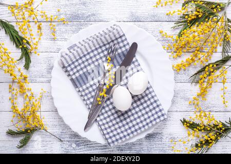 Easter table setting. White eggs, linen napkin, white plate, mimosa flowers and cutlery on a wooden table. Top view Stock Photo