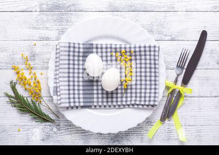Easter table setting. White eggs, linen napkin, white plate, mimosa flowers and cutlery on a wooden table. Top view Stock Photo