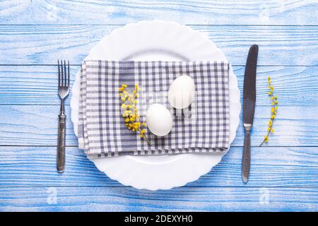 Easter table setting. White eggs, napkin on a plate, mimosa flowers, fork, knife on a blue wooden table. View from above Stock Photo