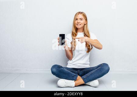 A beautiful young girl wearing a white t-shirt is sitting on the floor with her legs crossed showing a blank mobile phone screen on a grey background Stock Photo