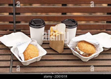Fast food. Paper cups with coffee, hamburger boxes and fries on a park bench.Takeaway food concept. Stock Photo