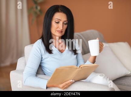 Mature lady reading book and holding cup of coffee Stock Photo