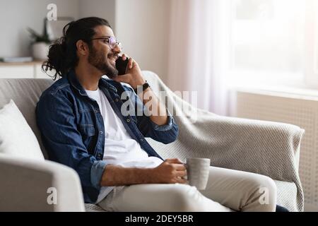 Smiling Indian Guy Resting At Home, Talking On Cellphone And Drinking Coffee Stock Photo