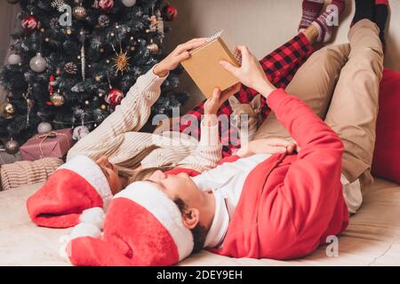 Romantic couple exchanging gifts near decorated christmas tree. Man giving christmas present surprise to smiling woman in sweater while relaxing on Stock Photo