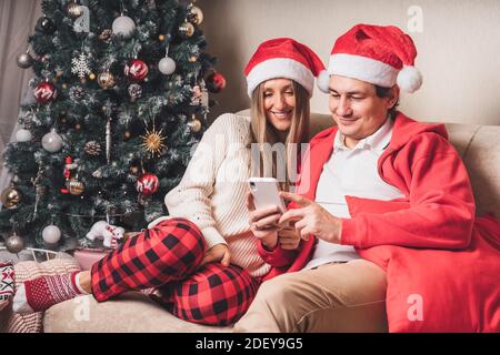 Portrait of lovely couple husband and wife in red Santa hat having a video call or taking selfie with their smartphone enjoy christmas time at home. Stock Photo
